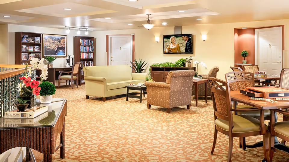Bright, furnished senior living common lounge with sofas, armchairs, tables, bookshelves, plants, and a wall-mounted TV.