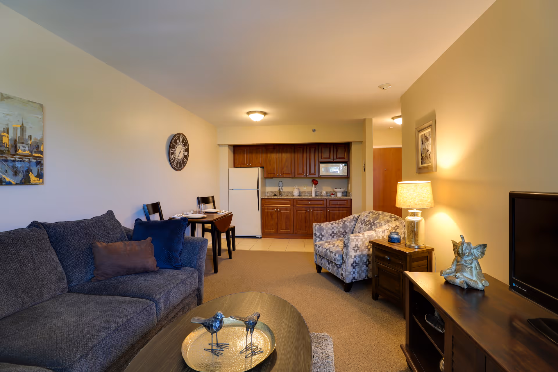 Cozy living room with a sofa, patterned armchair, coffee table and TV stand, with a small kitchenette and dining table visible in the background.