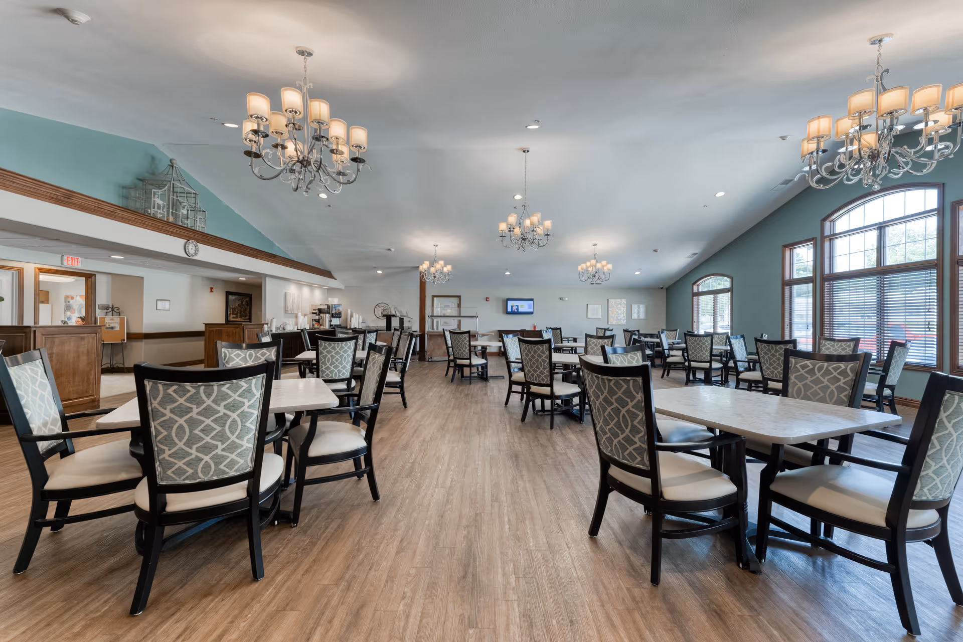 Spacious dining room with multiple tables and chairs arranged neatly on a wooden floor. The room features large windows with blinds, light blue walls, and several elegant chandeliers hanging from the ceiling. A counter area is visible in the background with coffee machines and other amenities.