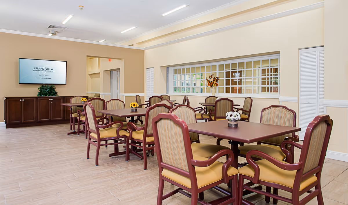 A dining room in a senior living community with several tables and chairs arranged neatly. Each table has a small flower arrangement. There is a large flat-screen TV mounted on the wall displaying the Grand Villa Senior Living Community logo. The room has beige walls, a tiled floor, and a window with a grid design letting in natural light.
