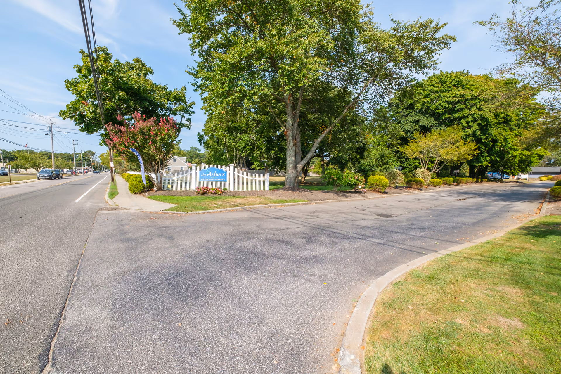 Entrance to The Arbors assisted living community with a white picket fence, a blue sign, and surrounding trees and greenery along a paved road on a sunny day.