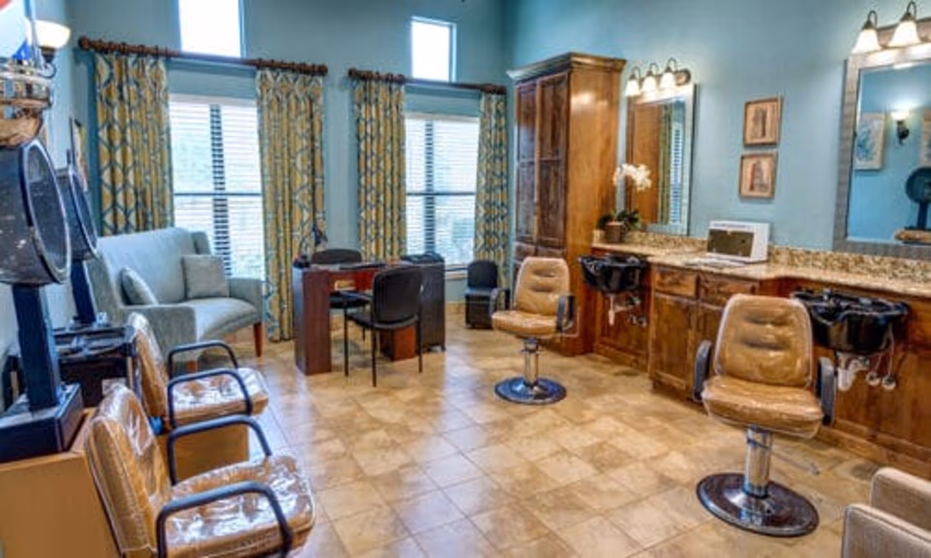 Interior of a salon room with two brown salon chairs in front of sinks and mirrors, a wooden cabinet, a small desk with chairs, a light blue loveseat, and three large windows with patterned curtains.