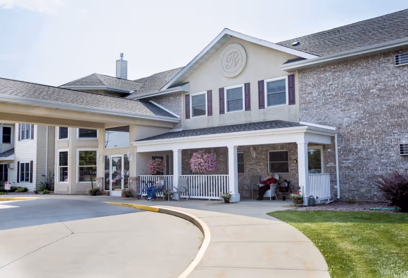 Exterior view of Renaissance by Rennes - Wisconsin Rapids senior living facility showing a covered entrance with a curved driveway. Two elderly individuals are seated on the porch under a roof supported by white columns. The building features beige siding, brick walls, and multiple windows with purple shutters. Hanging flower baskets and potted plants decorate the porch area.