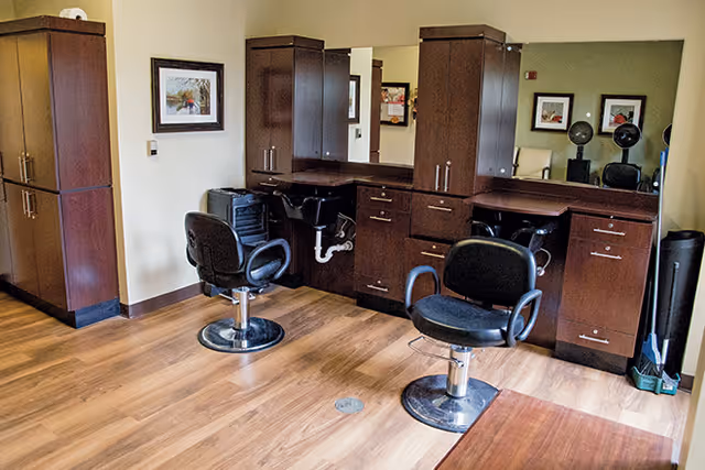 Interior view of a salon area with two black salon chairs in front of dark wooden cabinets and mirrors. The floor is wooden, and there are framed pictures on the walls.