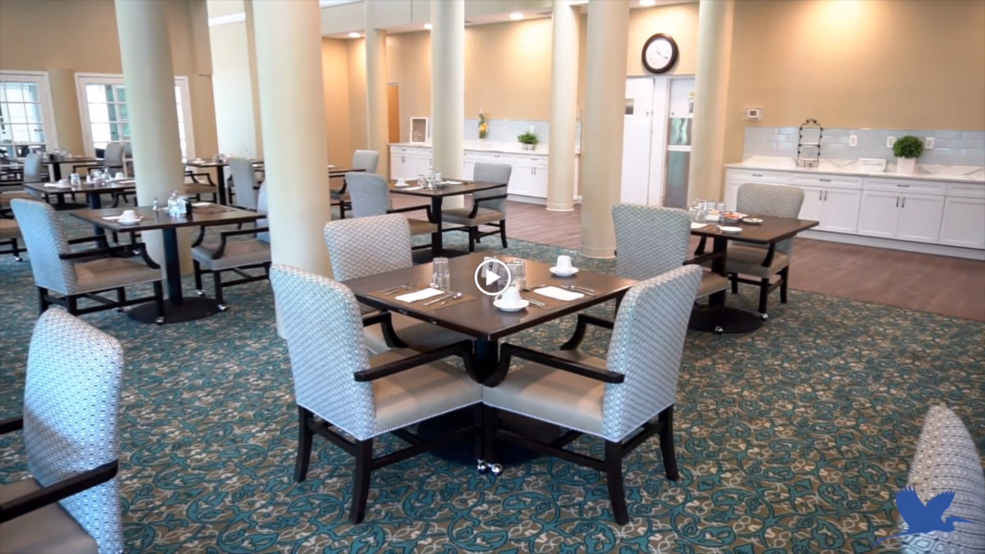 Dining room with multiple set tables and upholstered chairs around decorative columns and patterned carpet.