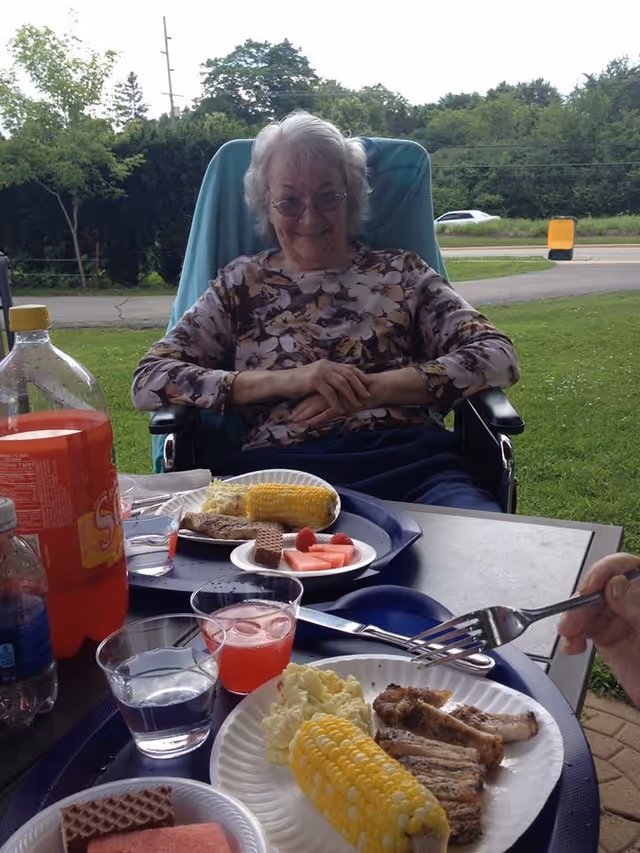 An elderly woman sitting outdoors in a wheelchair at a table with plates of food including corn on the cob, potato salad, grilled meat, watermelon slices, and wafer cookies. There are also drinks on the table including a bottle of orange soda, a glass of water, and a glass of pink beverage. The background shows green grass, trees, and a road with a car passing by.