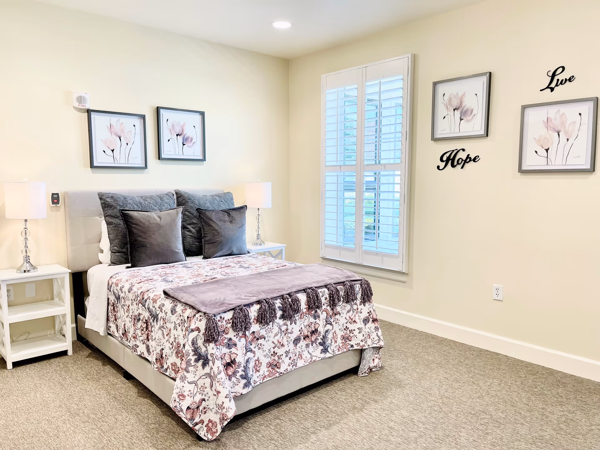 A neatly made bed with a floral patterned bedspread and multiple pillows in a softly lit bedroom. Two white nightstands with lamps flank the bed. The walls are decorated with framed floral artwork and the words 'Live' and 'Hope'. A window with white plantation shutters lets in natural light.