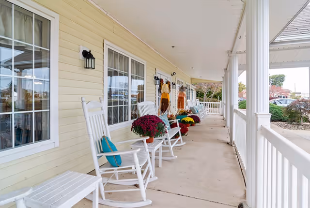 A covered porch area with white rocking chairs and small tables, decorated with colorful potted flowers. The porch has cream-colored siding, large windows, and white railings, overlooking a landscaped outdoor area with shrubs and trees.