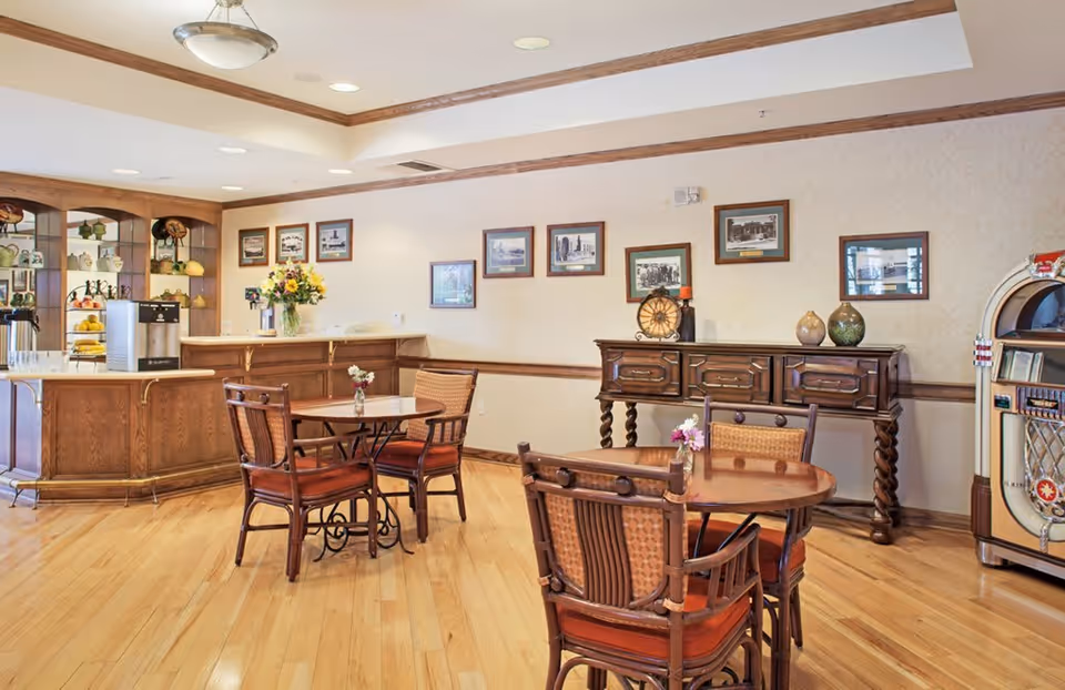 A cozy dining area with wooden floors and furniture, including two round tables each surrounded by four chairs. A wooden counter with a coffee machine and a vase of flowers is visible in the background, along with framed pictures on the wall and a vintage jukebox on the right side.
