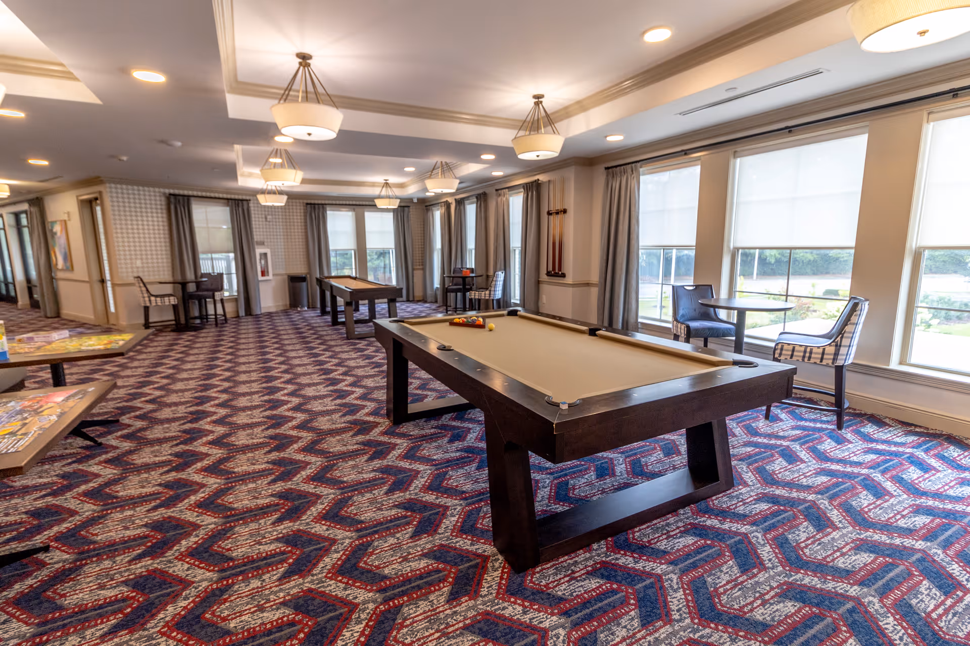 A spacious game room with patterned carpet featuring a pool table in the center and a shuffleboard table in the background. The room has large windows with curtains allowing natural light to fill the space. There are also small tables with chairs near the windows and along the walls, and ceiling lights provide additional illumination.