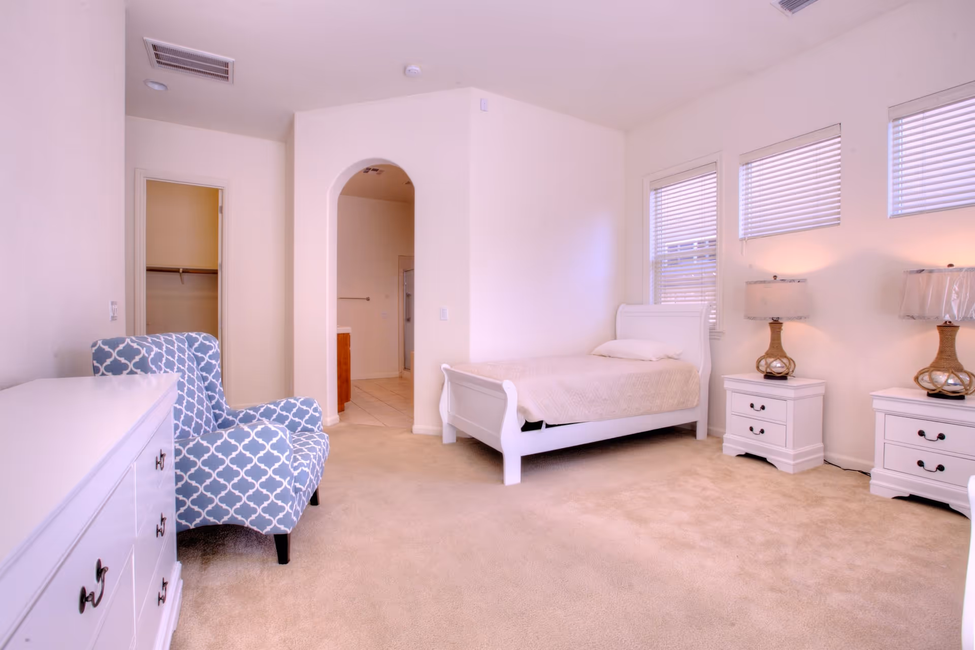 A bright bedroom with beige carpet, a white bed with a beige quilt, two white nightstands each with a lamp, a blue patterned armchair, and a white dresser. There is an arched doorway leading to a bathroom and a closet door visible.