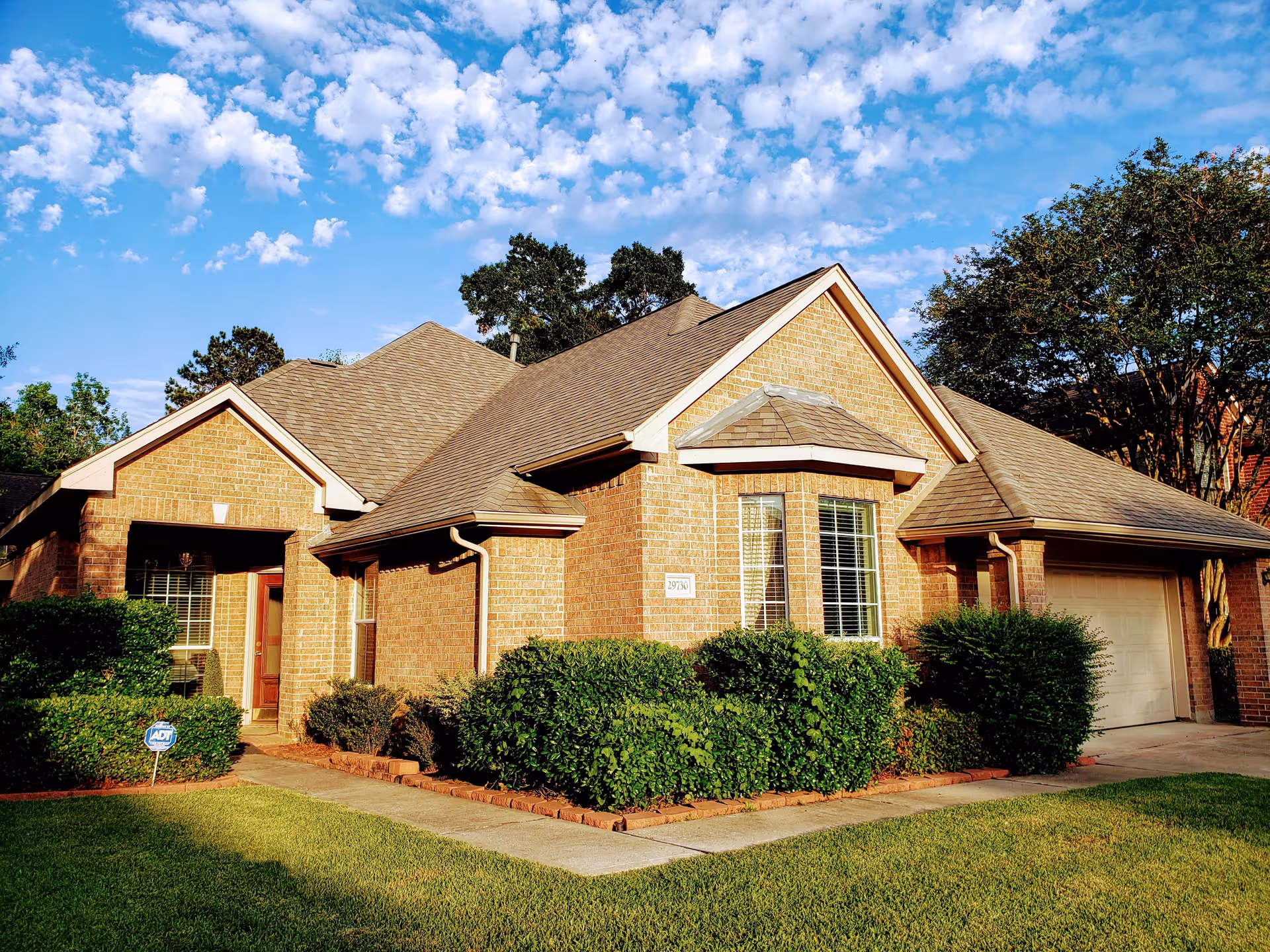 Exterior view of a single-story brick house with a well-maintained lawn, shrubs, and a driveway leading to a garage under a partly cloudy blue sky.