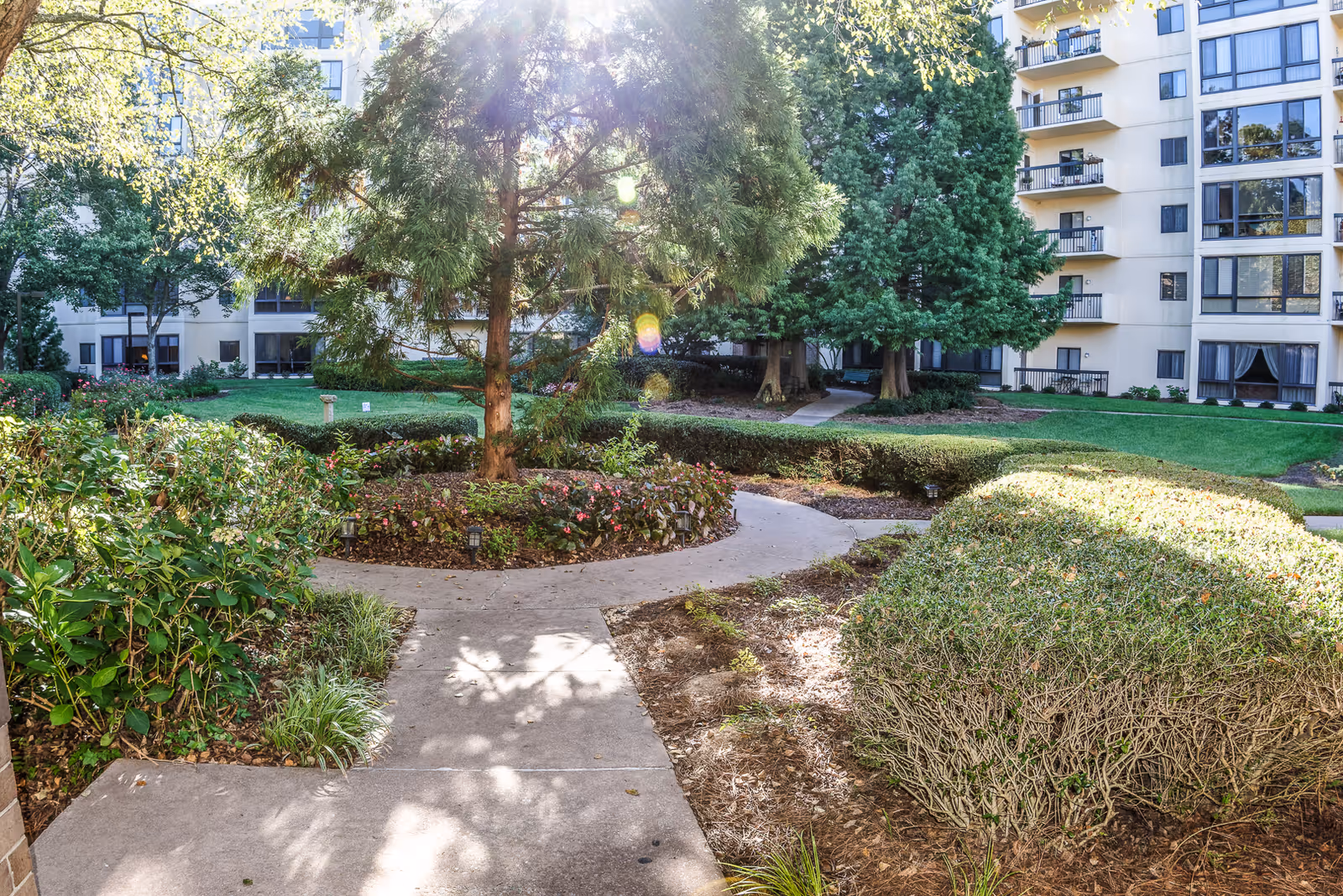 Sunlit outdoor garden area with a concrete pathway winding through trimmed bushes, trees, and flower beds, surrounded by a multi-story residential building with balconies and large windows.