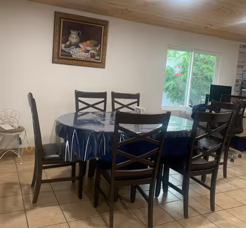 A dining area with a round table covered by a dark blue plastic tablecloth and surrounded by six black wooden chairs. There is a window showing greenery outside, a framed painting of fruit on the wall, and a computer desk with a monitor and chair in the background.