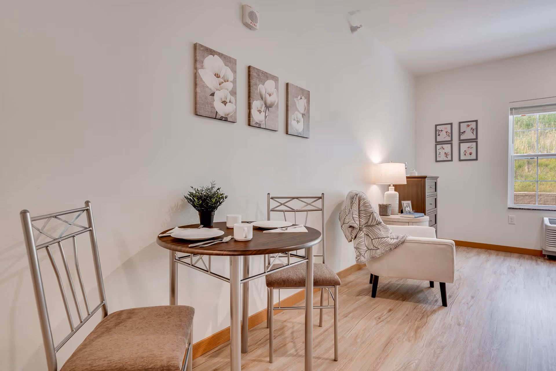 Bright furnished living/dining area in a senior apartment with a small round dining table, metal chairs, an armchair with a blanket, lamp, and wall art.