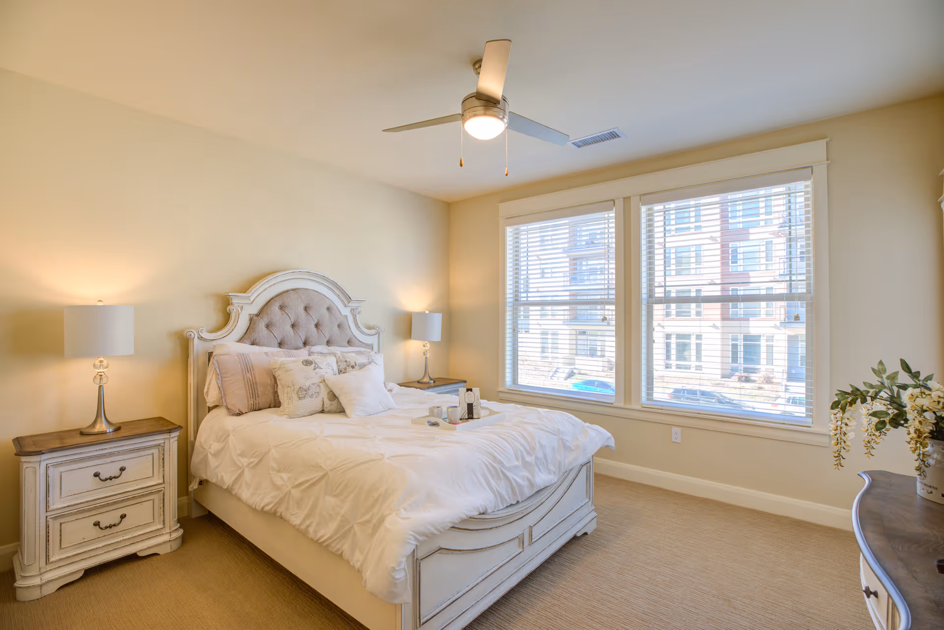 Sunlit bedroom featuring a white upholstered bed with pillows, matching nightstands and lamps, large windows with blinds, and a ceiling fan.