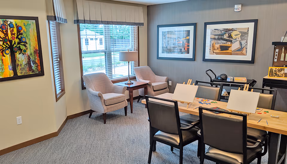 Bright senior living community activity room with a table set for painting, chairs and easels, two armchairs by a window, and framed artwork on the walls.