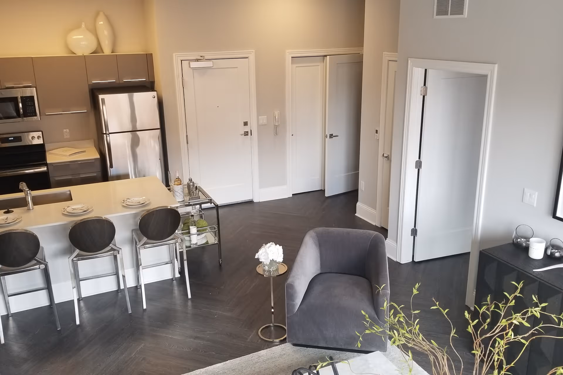 Modern open-concept living space featuring a kitchen with stainless steel appliances, a breakfast bar with three chairs, a gray armchair, a small round side table with a flower vase, and a dark wood floor. There are multiple white doors leading to other rooms and a black cabinet with decorative items on top.