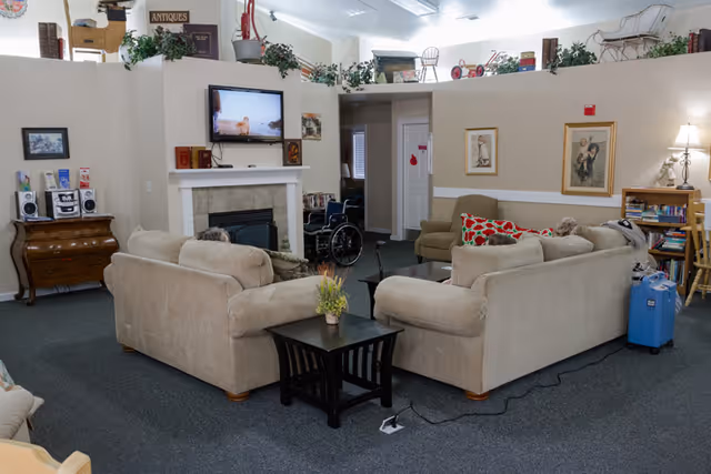 A cozy living room area in a memory care facility with beige sofas arranged around a small black coffee table. A flat-screen TV is mounted above a fireplace, and there are various decorations including framed pictures, plants, and bookshelves. A wheelchair is visible near the back, and the room has a carpeted floor with soft lighting.