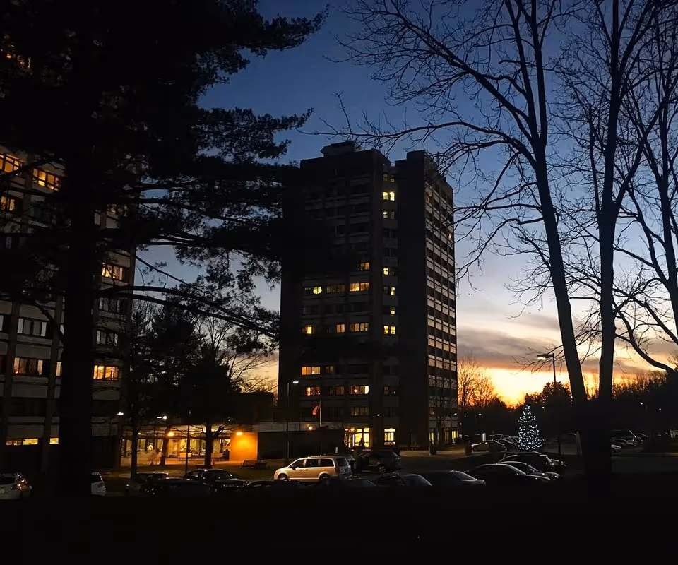 Exterior view of a tall apartment building at dusk with lights on in some windows. Trees without leaves and a decorated Christmas tree are visible in the foreground along with parked cars. The sky shows a sunset with a gradient from orange near the horizon to dark blue higher up.