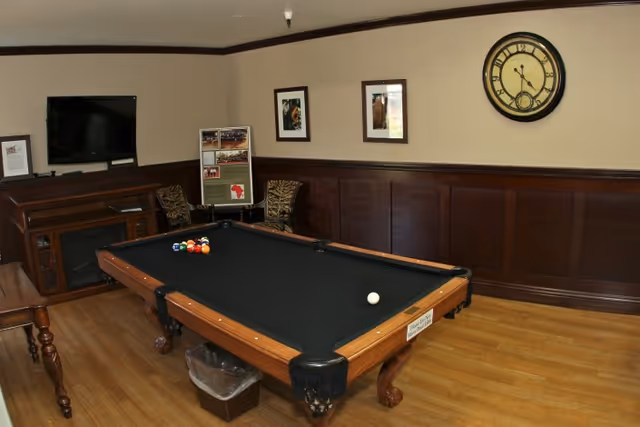 A room with a pool table in the center, a television mounted on the wall above a wooden cabinet, two chairs with a small table between them, framed pictures on the wall, and a large clock. The floor is wooden and the walls have dark wood paneling on the lower half.