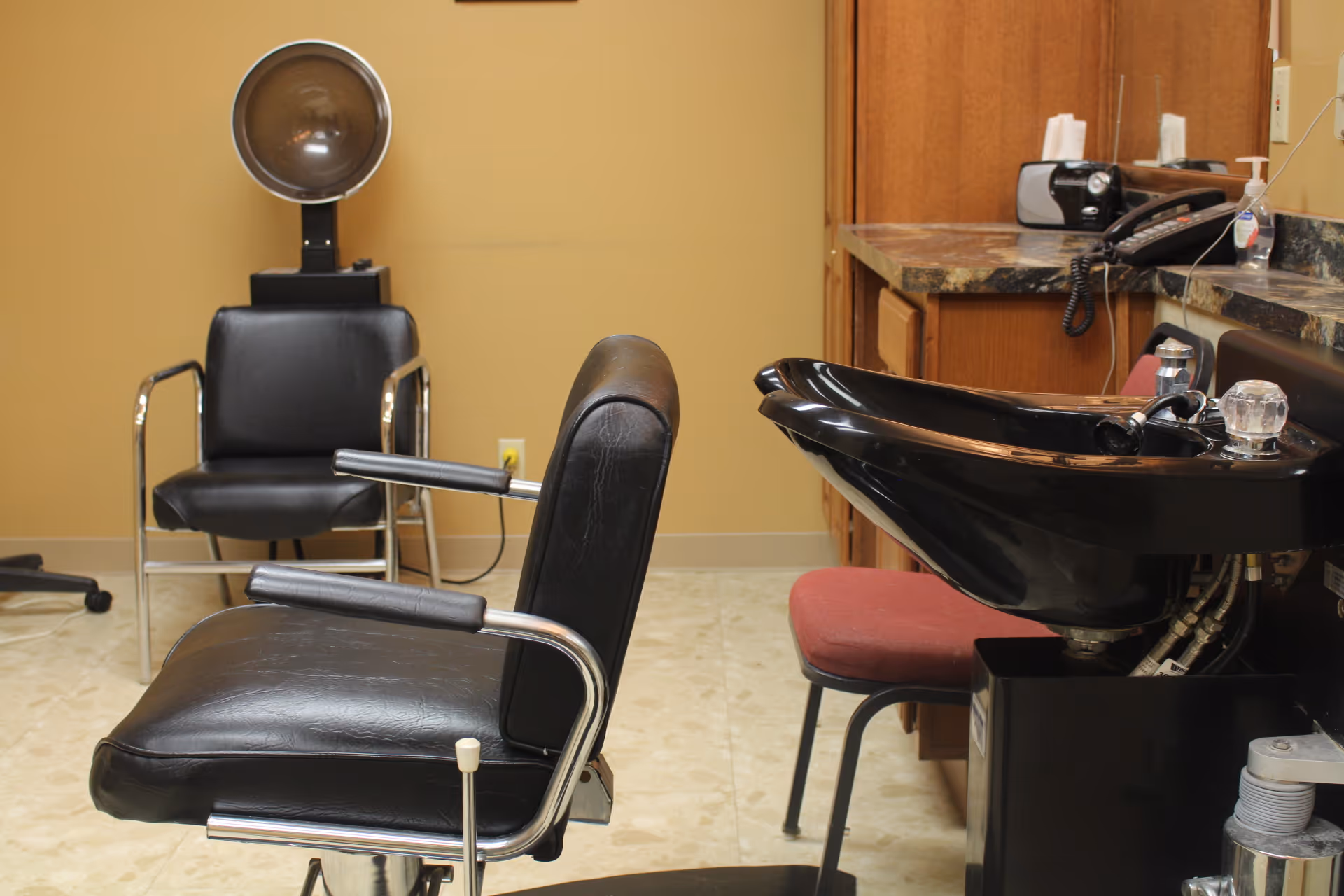 Interior of a salon area in a nursing home featuring a black salon chair, a hair washing sink with a red chair in front of it, a hair dryer chair in the background, and a countertop with a telephone and other items.