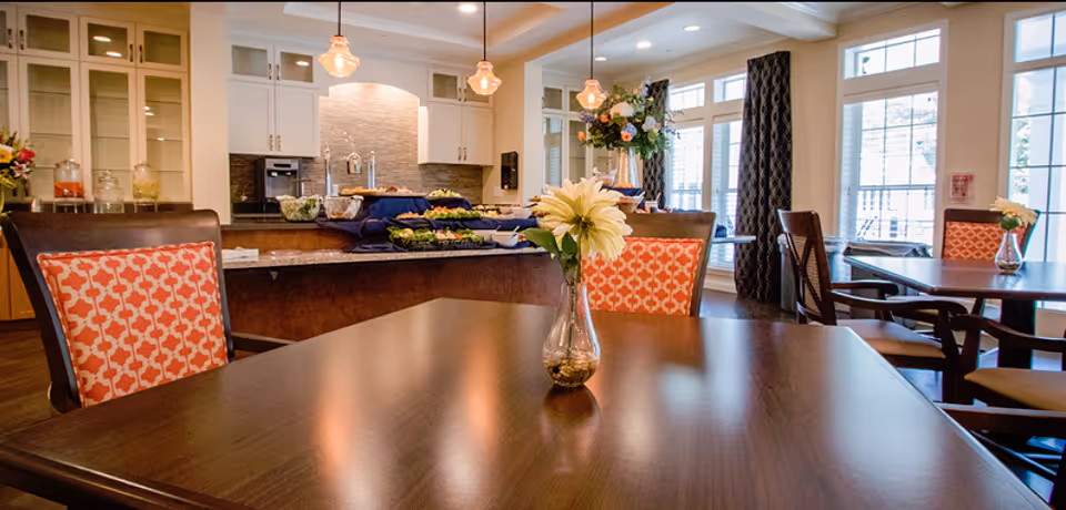 Dining room with wooden tables and patterned upholstered chairs, a central buffet counter, pendant lights, and large windows.