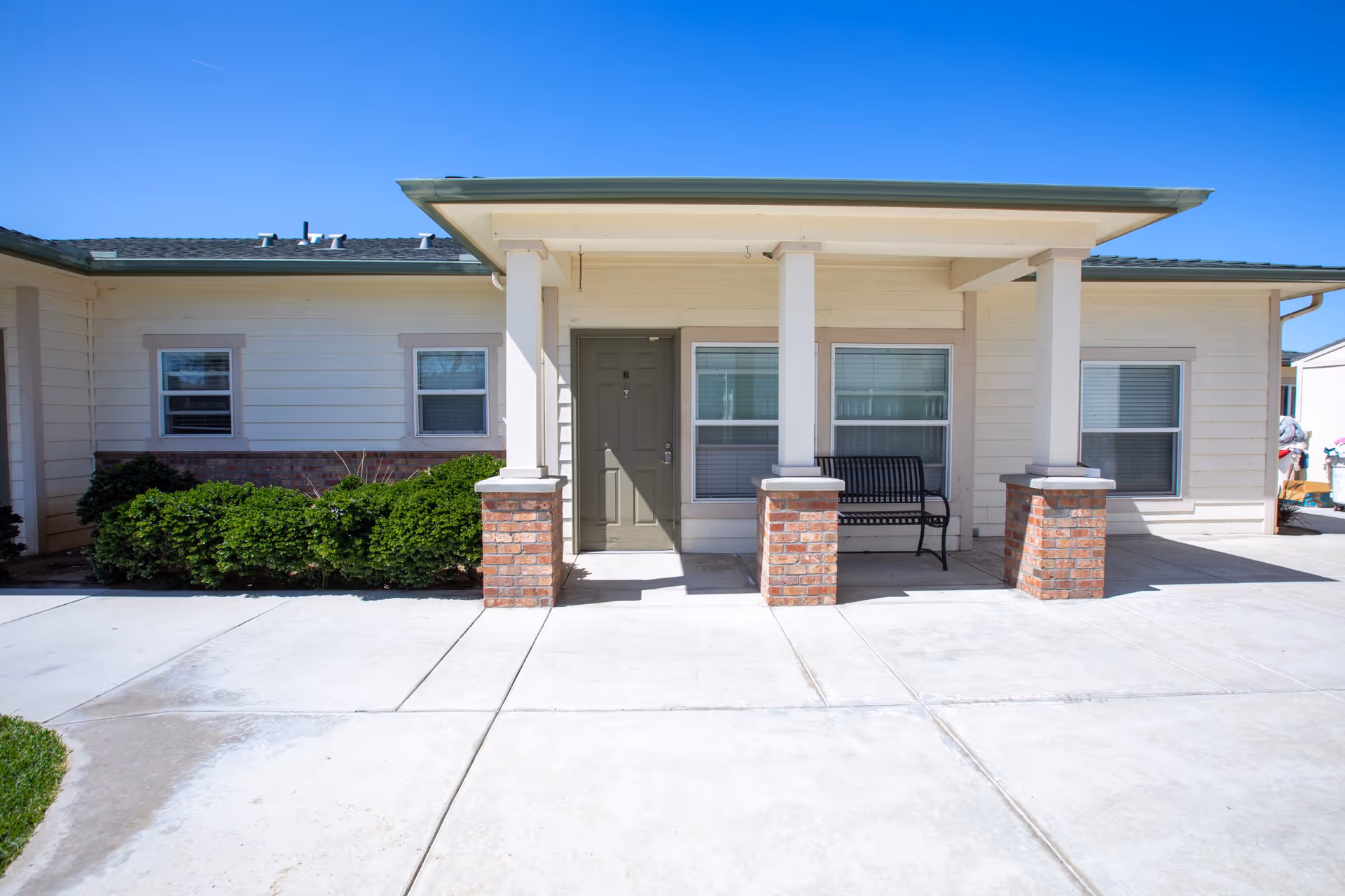 Front exterior view of a single-story senior living facility unit with a covered porch supported by four white columns with brick bases. The porch has a black bench, and there are three windows with white frames. The building has light-colored siding and a green roof. The concrete walkway and driveway are visible in the foreground under a clear blue sky.