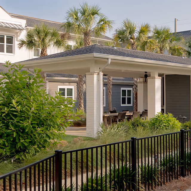 Outdoor covered seating area with a roof supported by white brick columns, surrounded by palm trees and greenery, with a black metal fence in the foreground and residential buildings in the background.