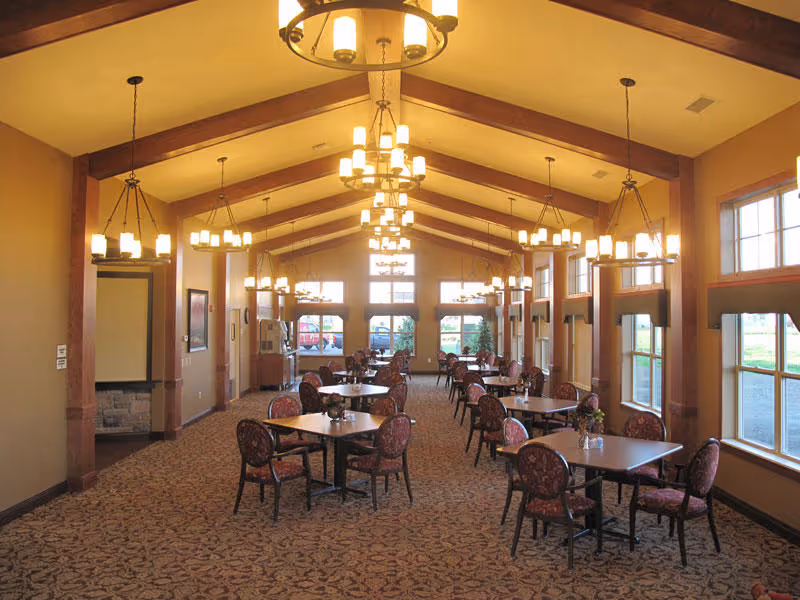 A spacious dining room with multiple square tables and cushioned chairs arranged in rows. The room features high vaulted ceilings with wooden beams and multiple hanging chandeliers providing warm lighting. Large windows line one side of the room, allowing natural light to enter and offering views of the outdoors. The carpet has a patterned design, and the walls are painted in a warm beige tone.