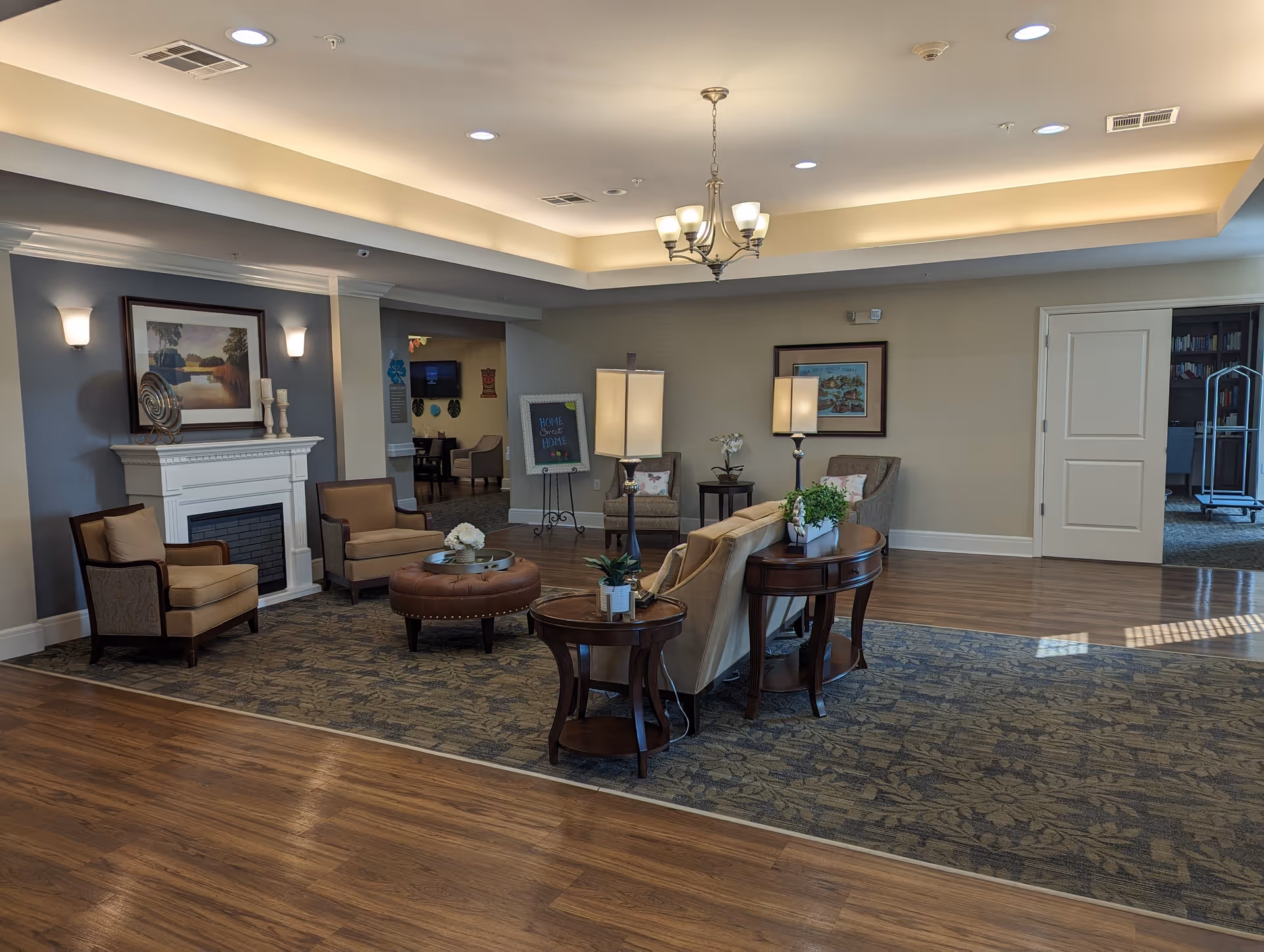 A spacious and well-lit living room area in a senior living facility with a beige sofa, two armchairs, a round ottoman, and wooden side tables with plants. The room features a white fireplace with decorative items and a framed landscape painting above it. There is a chandelier hanging from the ceiling and recessed lighting. The floor is a combination of wood and patterned carpet, and there are open doorways leading to other rooms.