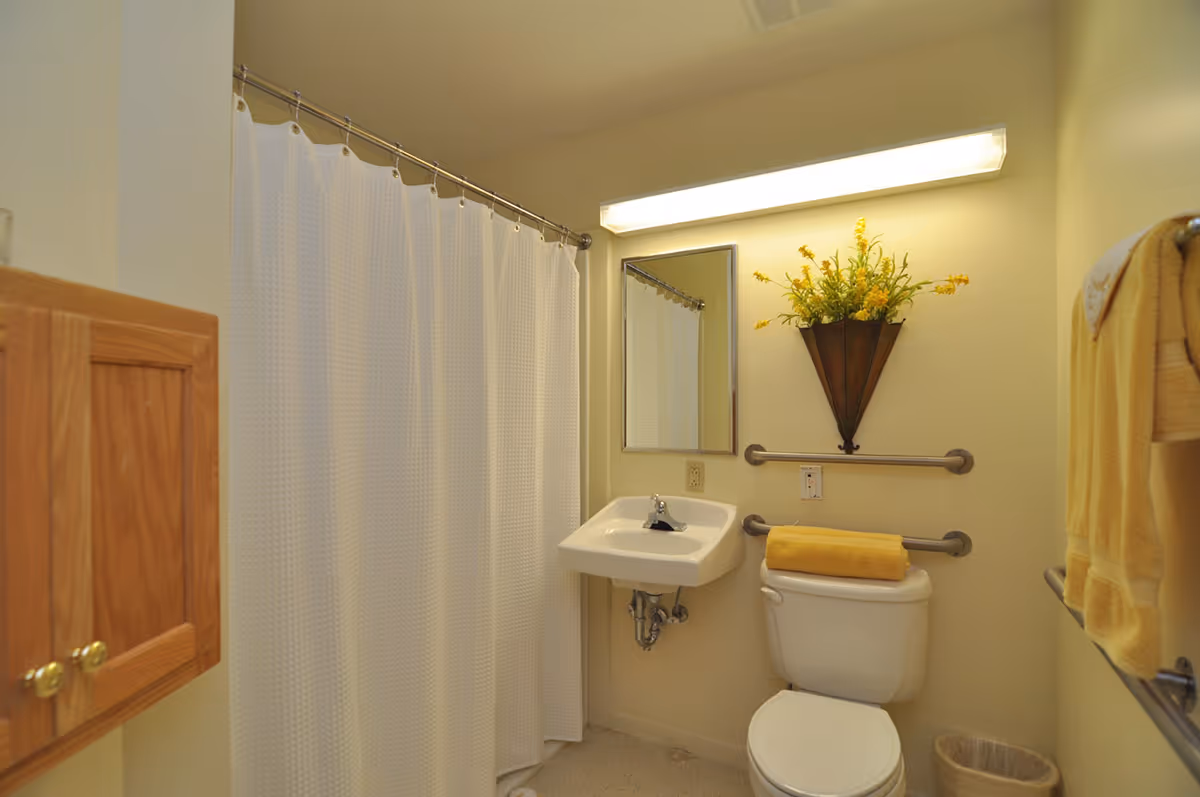A small bathroom with a white shower curtain, a wall-mounted sink, a toilet with a yellow towel on top, yellow towels hanging on a rack, a wall-mounted decorative flower arrangement, and grab bars for accessibility.