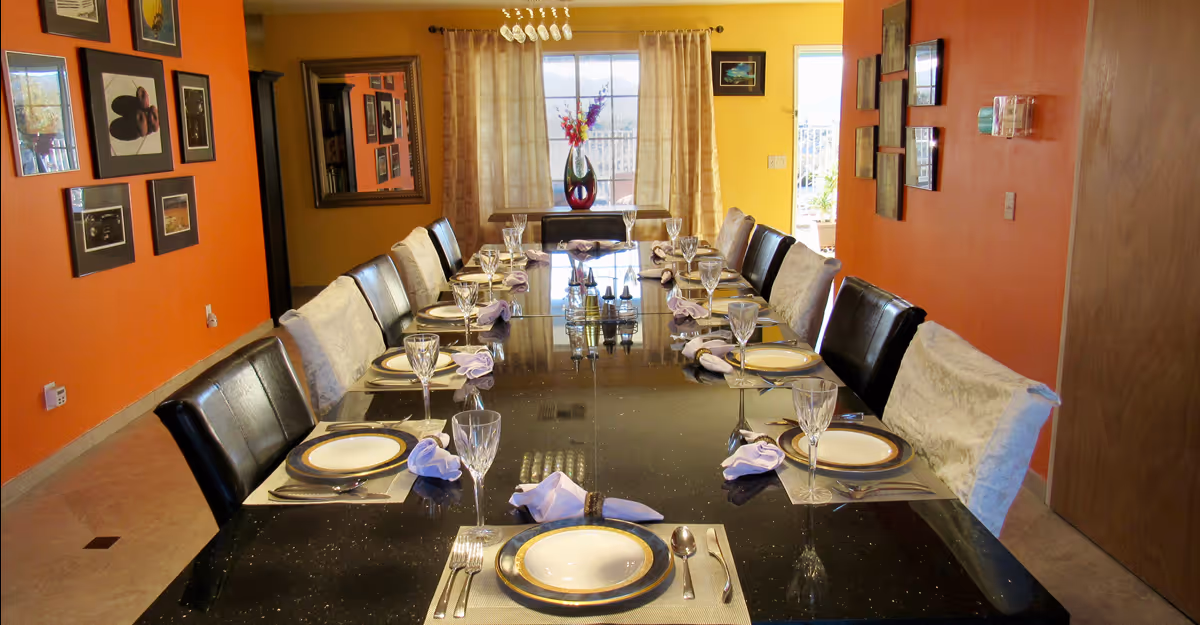 A long dining table set for a meal with plates, glasses, silverware, and folded napkins in a room with orange and yellow walls. The room has framed pictures on the walls and a window with curtains at the far end, letting in natural light. There is a vase with flowers on a small table near the window.