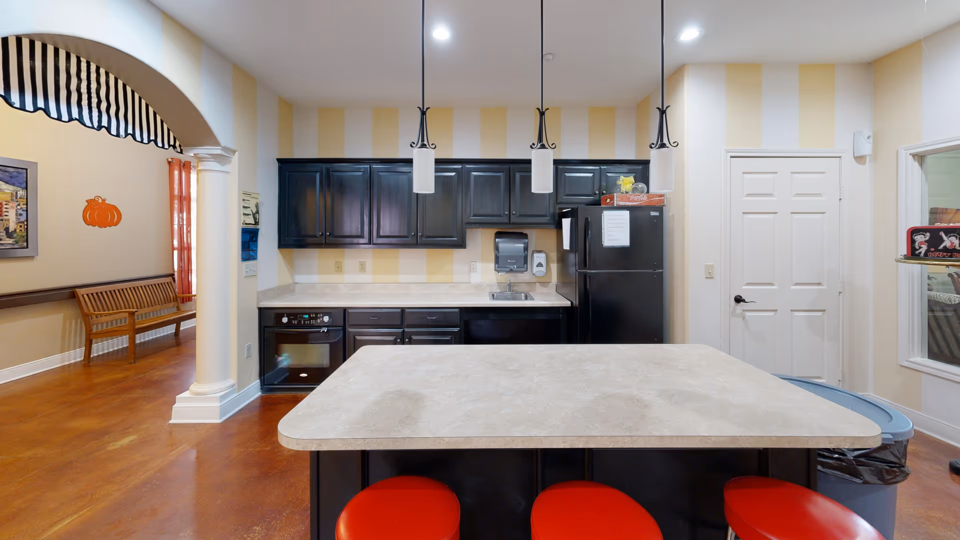 Interior view of a kitchen area with a large island countertop and three red stools. The kitchen has black cabinets, a black refrigerator, an oven, and a sink. The walls have yellow and white vertical stripes. To the left, there is an archway with a column leading to another room with a wooden bench and a pumpkin decoration on the wall.