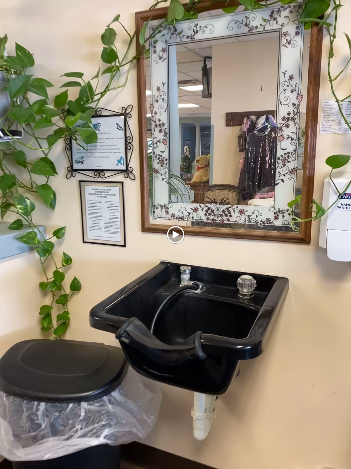 A black salon-style sink with a hose and a padded neck rest mounted on a beige wall. Above the sink is a decorative mirror with floral patterns on the frame. To the left of the mirror are two framed notices on the wall, and a green leafy plant drapes around the area. A black trash can with a plastic liner is positioned below the sink. On the right side of the image, a wall-mounted hand sanitizer dispenser is visible.