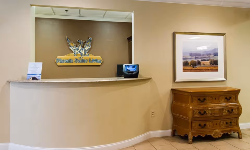 Reception area with a curved beige counter, a sign on the wall behind reading 'Phoenix Senior Living' with a phoenix logo, a small digital device on the counter, and a wooden chest of drawers with a framed landscape painting above it on the right side.