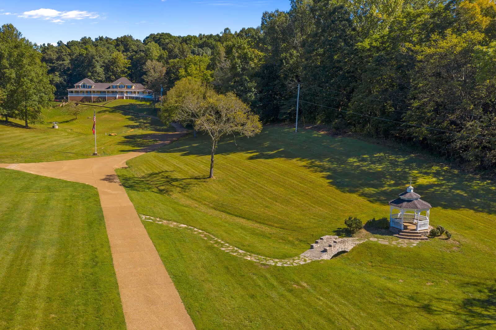 A large green lawn with a paved walkway leading to a building in the distance surrounded by trees. There is a small white gazebo on the right side of the lawn connected by a stone path, and an American flag on a flagpole near the center of the image.