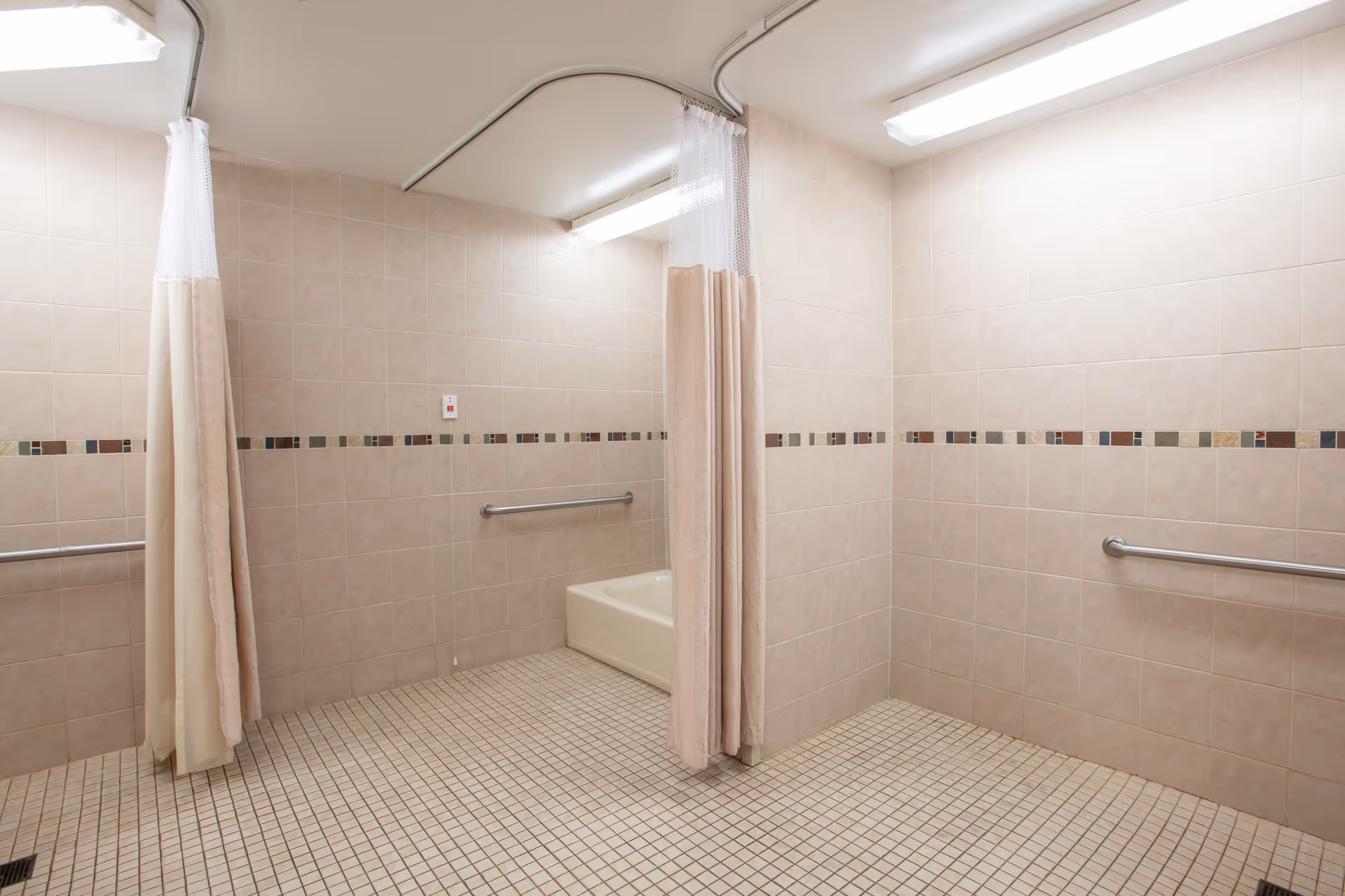 A tiled bathroom with beige walls and floor, featuring a bathtub, grab bars on the walls, and a shower curtain hanging from a ceiling-mounted rail. The room is well-lit with fluorescent ceiling lights.