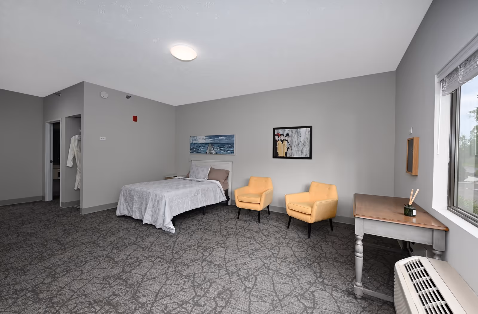 A senior living facility bedroom with a bed covered in a white quilt, two yellow armchairs, a wooden desk near a window, and two paintings on the gray walls. The room has carpeted flooring and an air conditioning unit below the window. A bathroom entrance and a closet with a white robe hanging are visible on the left side.