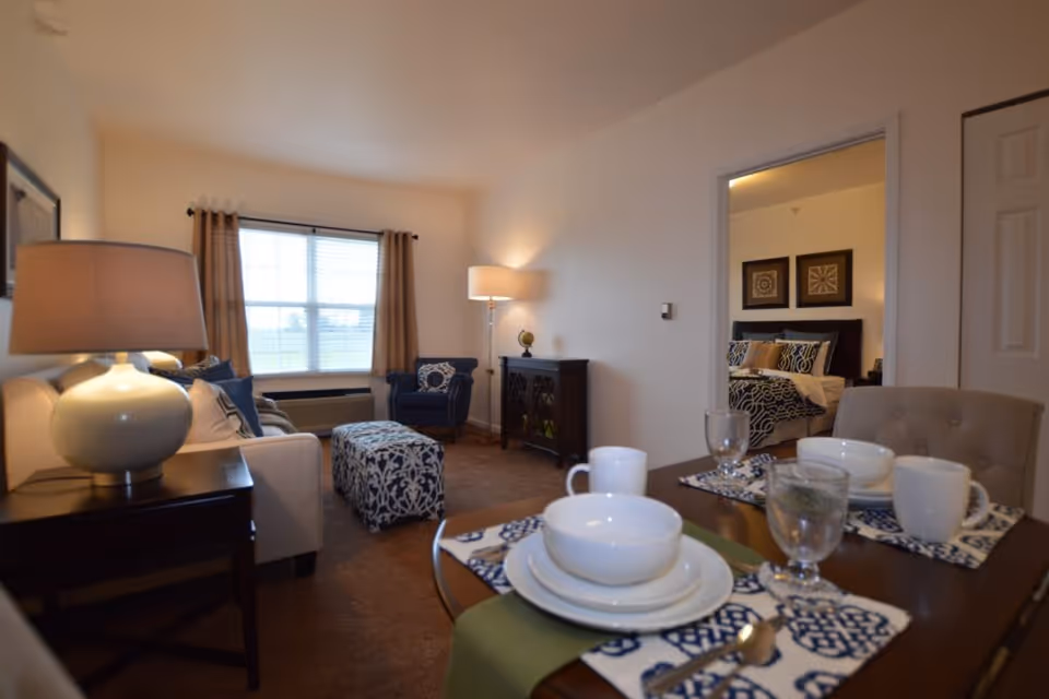Furnished living room and dining area with a set table in the foreground, sofa, armchair, ottoman, and a view into a bedroom.