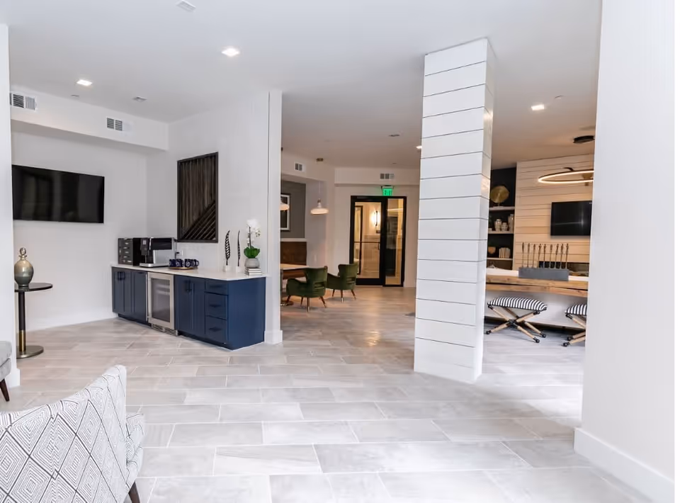 A modern interior common area with light gray tiled flooring, white walls, and recessed ceiling lights. There is a navy blue cabinet with a coffee machine and mugs on top, a wall-mounted TV, green upholstered chairs around a table in the background, and a white pillar with horizontal lines. The space appears bright and clean with a contemporary design.