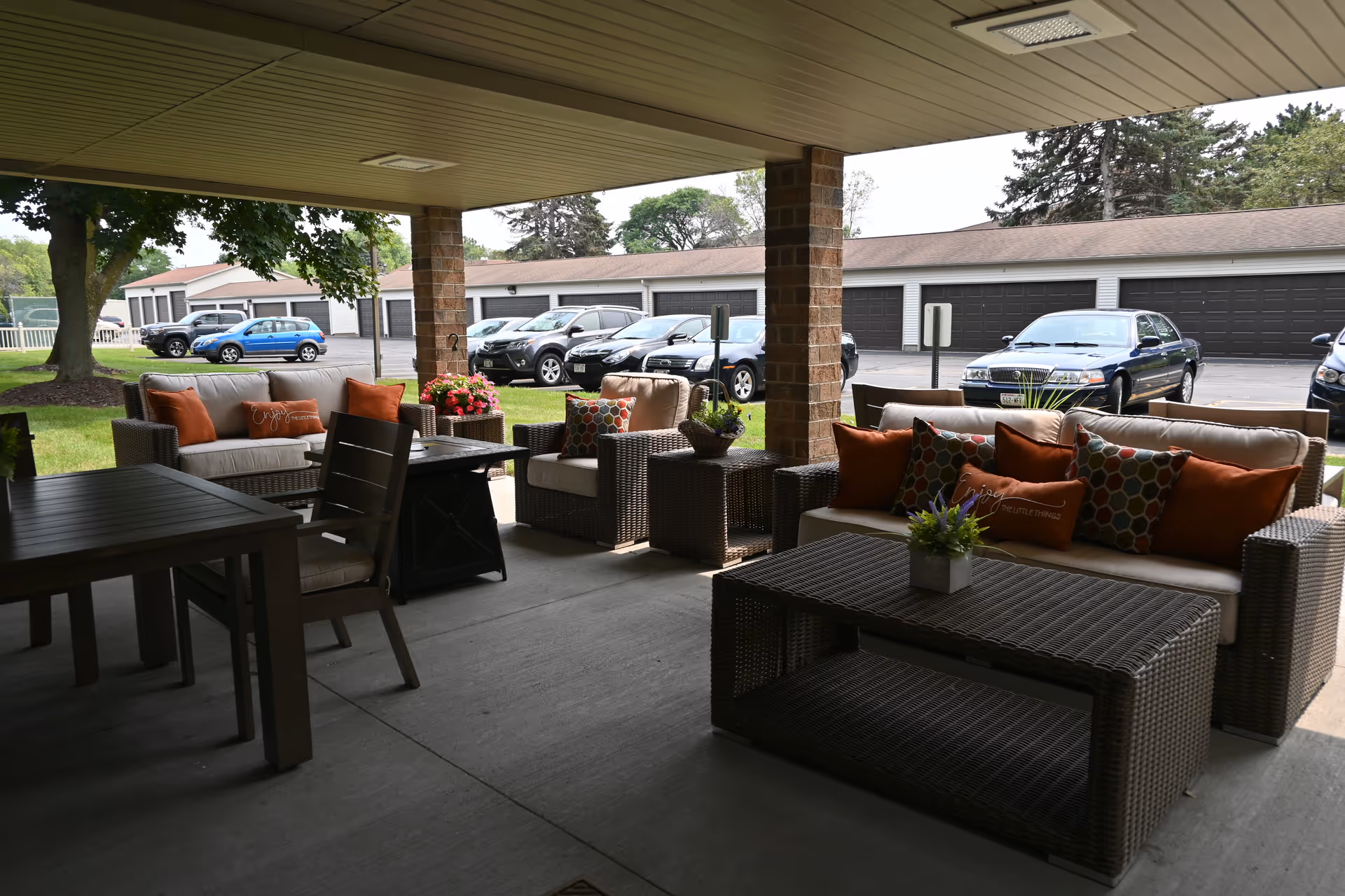 Covered outdoor patio area with wicker furniture including sofas and chairs with orange and patterned cushions, a coffee table, and a dining table with chairs. In the background, there is a parking lot with several parked cars and a row of garages. Trees and grass are visible beyond the patio.