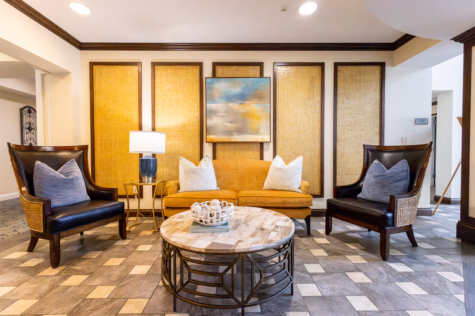 A cozy living room area featuring a mustard yellow sofa with two white pillows, flanked by two dark wooden armchairs with blue cushions. A round coffee table with a decorative bowl sits in front of the sofa. The wall behind has four large rectangular panels with a textured golden fabric and a colorful abstract painting in the center. The floor is tiled with a pattern of gray and beige tiles.
