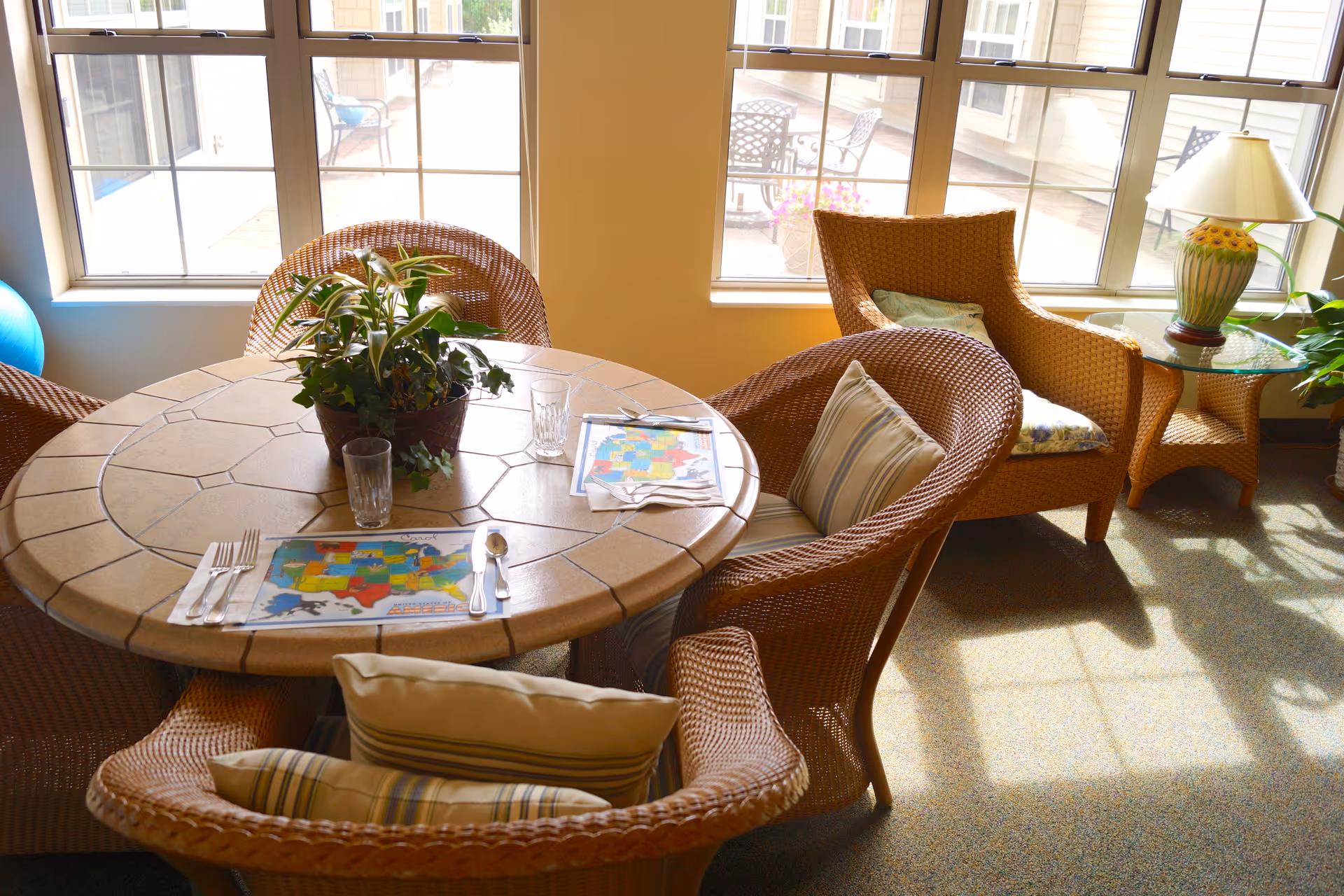 A sunlit dining area with a round tiled table surrounded by four wicker chairs with cushions. The table has placemats featuring a colorful map, utensils, and glasses. A potted plant is centered on the table. Large windows in the background show an outdoor patio with chairs and a table. A wicker armchair with a cushion and a glass-top side table with a decorative lamp are also visible.