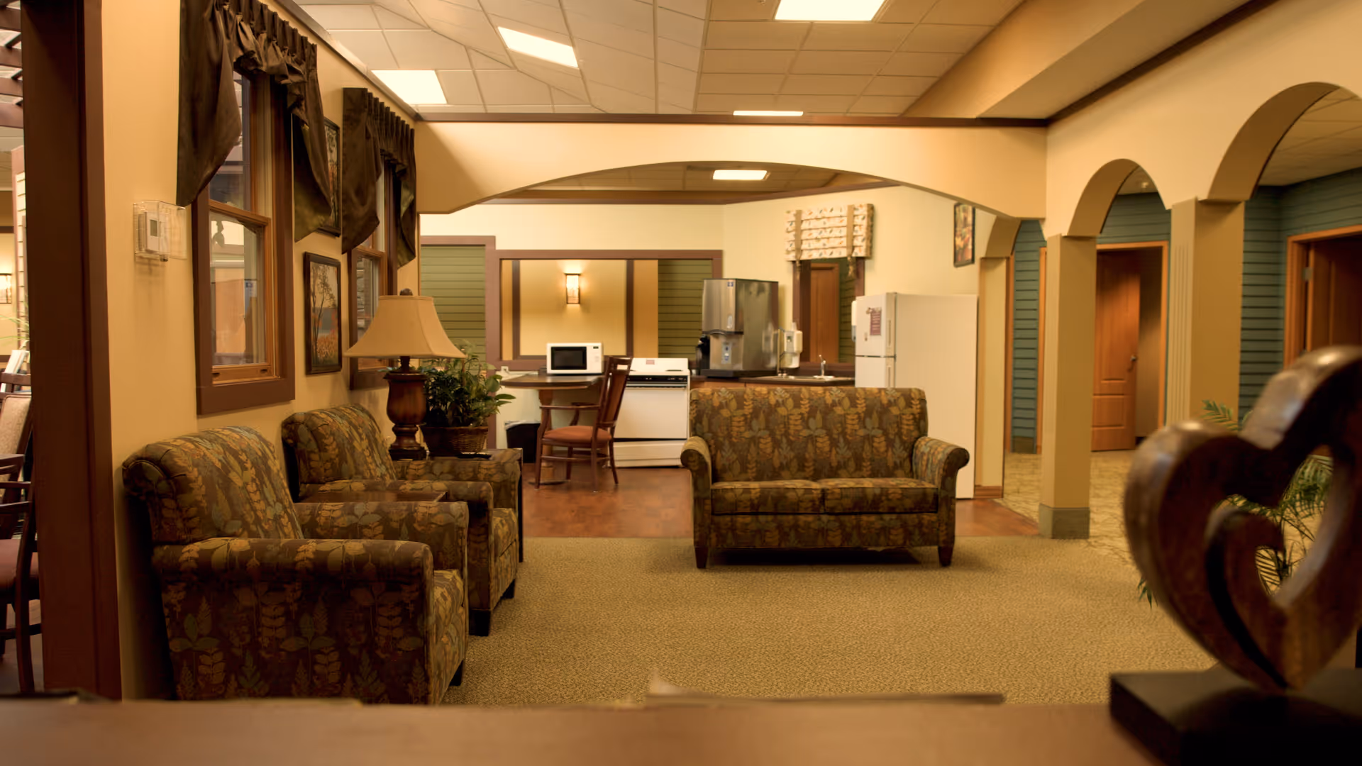Interior view of a common area in a senior living facility featuring patterned armchairs and a loveseat, a lamp on a side table, a small kitchen area with a refrigerator, microwave, and coffee machine, and arched doorways leading to other rooms.
