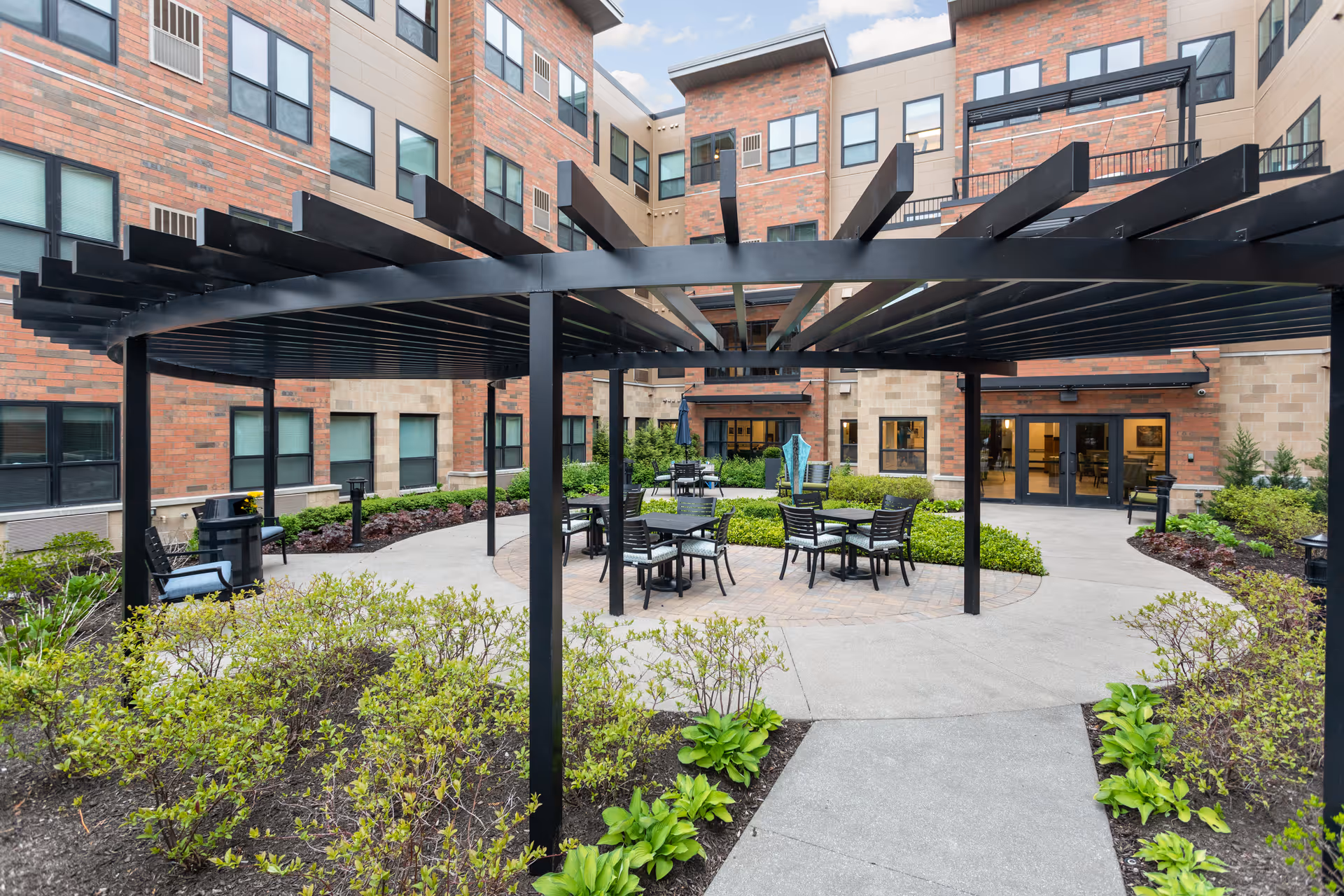 Courtyard with a circular black pergola, outdoor tables and chairs, and landscaping in front of a multi-story brick senior living building.