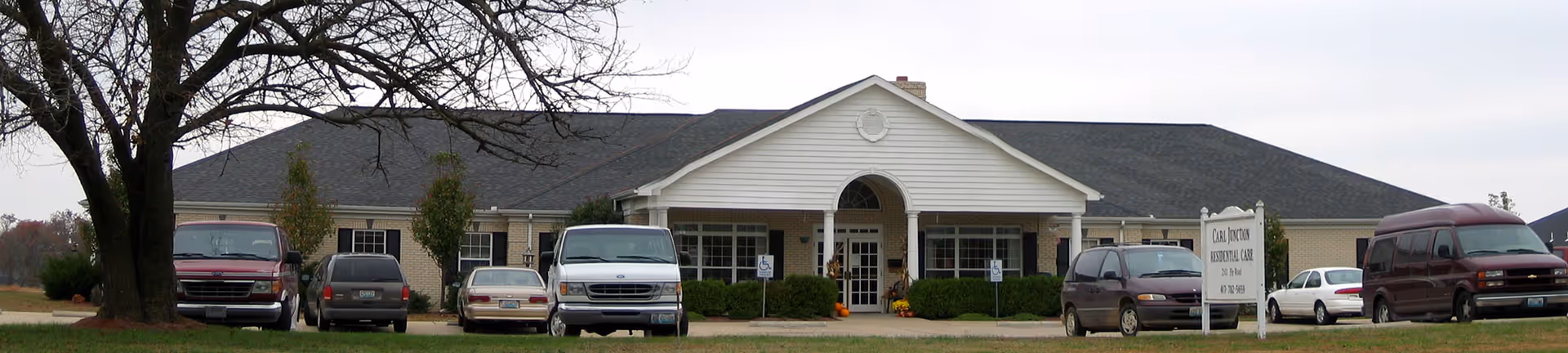 Front exterior view of Carl Junction Residential Care Center, a single-story building with a white entrance porch and several parked vehicles in front. There are trees and bushes around the building under an overcast sky.