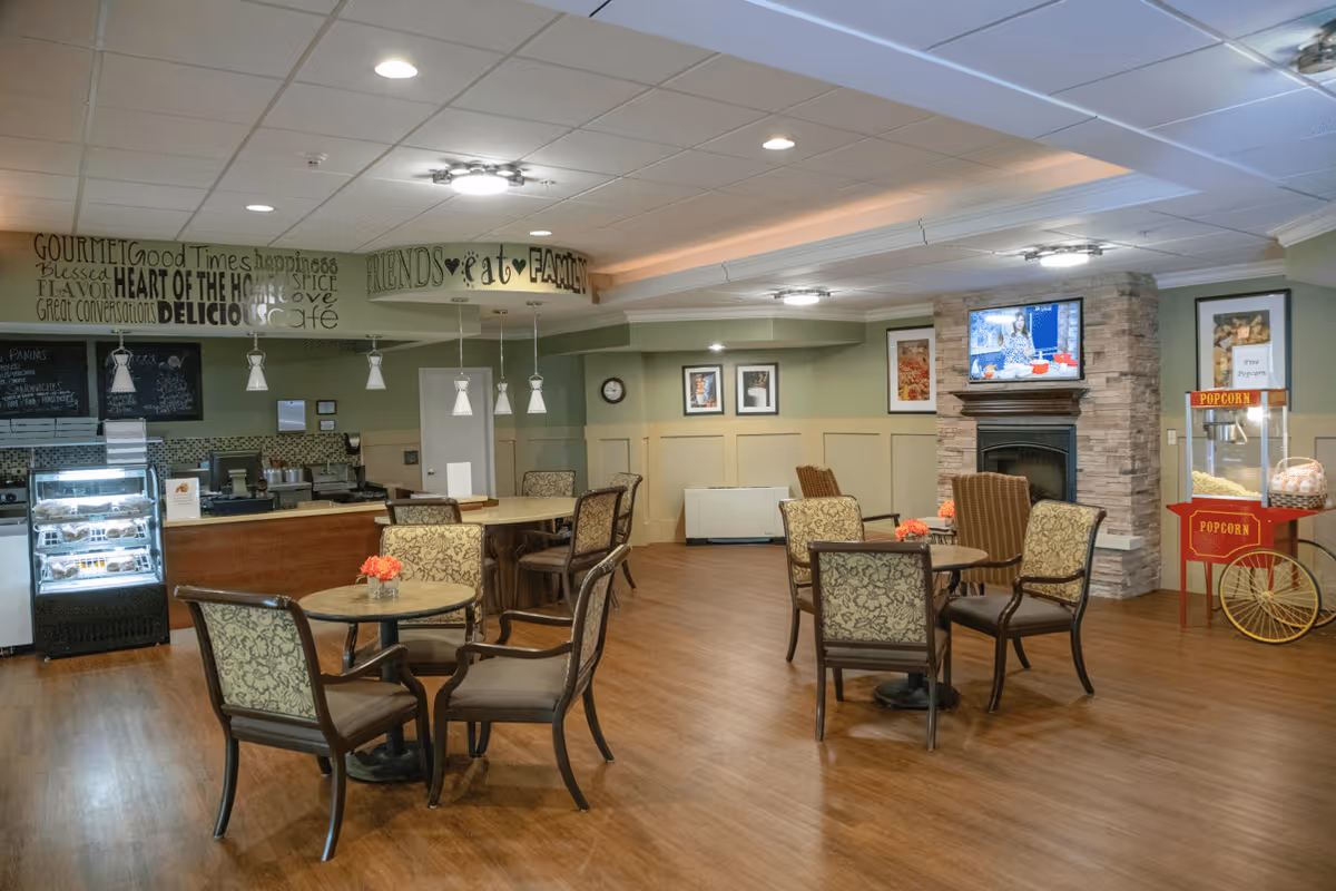 A cozy common area in a retirement community featuring several round tables with cushioned chairs, a popcorn machine, a stone fireplace with a mounted TV above it, and a small café counter with a display case of pastries. The walls are decorated with framed pictures and positive words like 'Friends', 'Family', and 'Heart of the Home'.