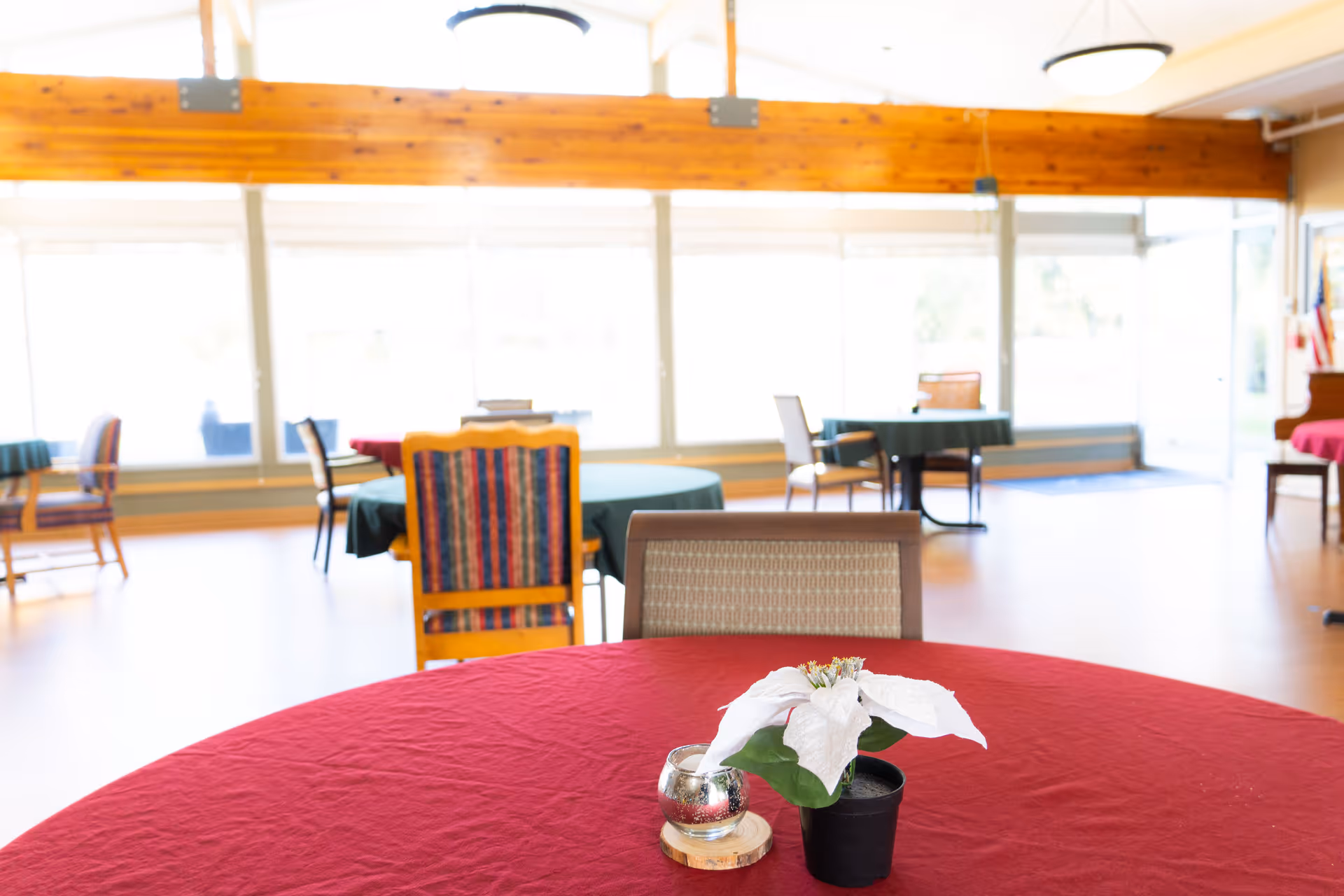 A bright dining area with round tables covered in red and green tablecloths. Each table has chairs around it, and the table in the foreground has a small potted white poinsettia and a silver candle holder. Large windows let in natural light, and wooden beams are visible on the ceiling.