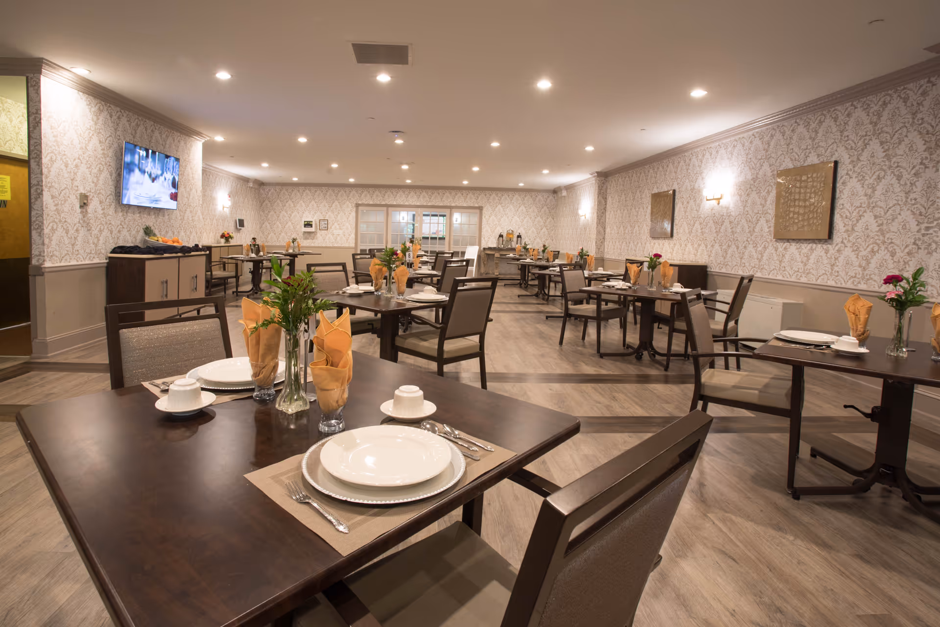 A spacious dining room with multiple dark wood tables and chairs arranged neatly. Each table is set with white plates, cups, silverware, and folded orange napkins. There are small flower vases on the tables. The room has patterned wallpaper, wooden flooring, and soft lighting from ceiling lights and wall sconces. A television is mounted on the left wall.